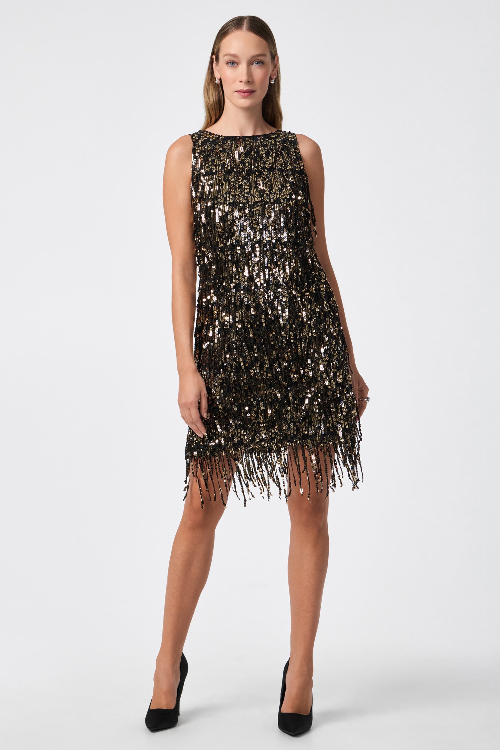 A woman wears the Nini Dress, a sleeveless, knee-length black dress with gold and black sequins and fringe, paired with black high heels, standing against a plain white background.