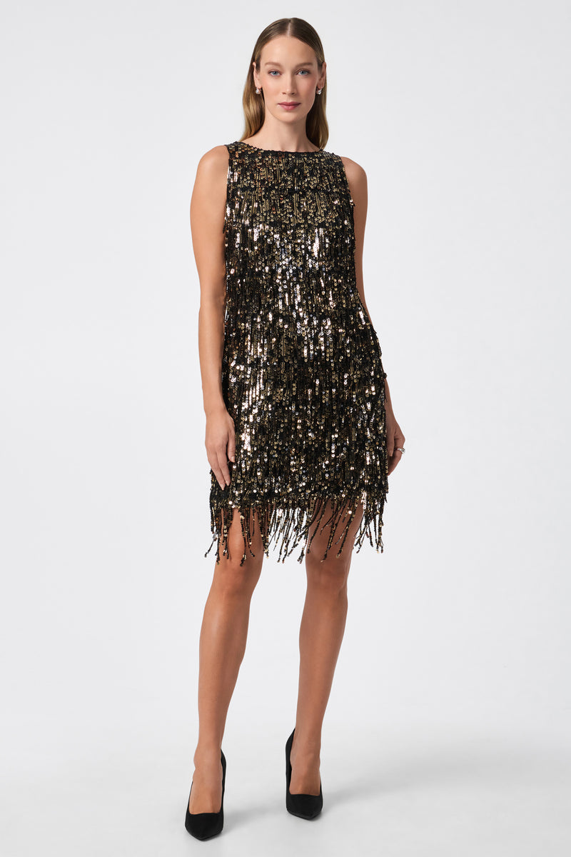 A woman stands against a white background wearing the Nini Dress in BLACK/GOLD, a sleeveless, knee-length dress with shimmering sequins and fringe, paired with black high heels.