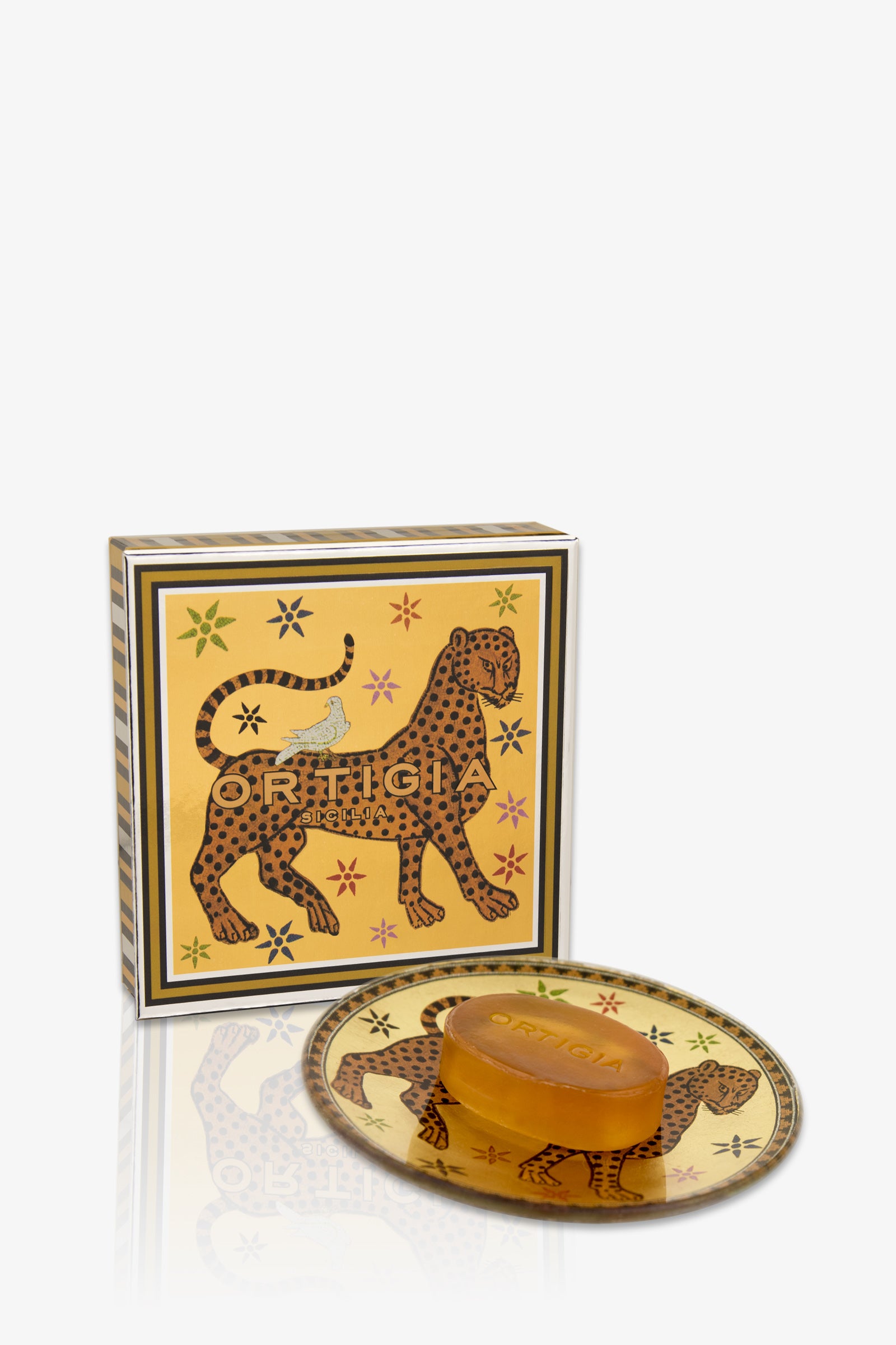 The Ortigia Ambra Nera Soap/Plate set features an amber soap bar and gold plate with Ortigia branding. The packaging showcases a leopard design and colorful stars on a gold background.