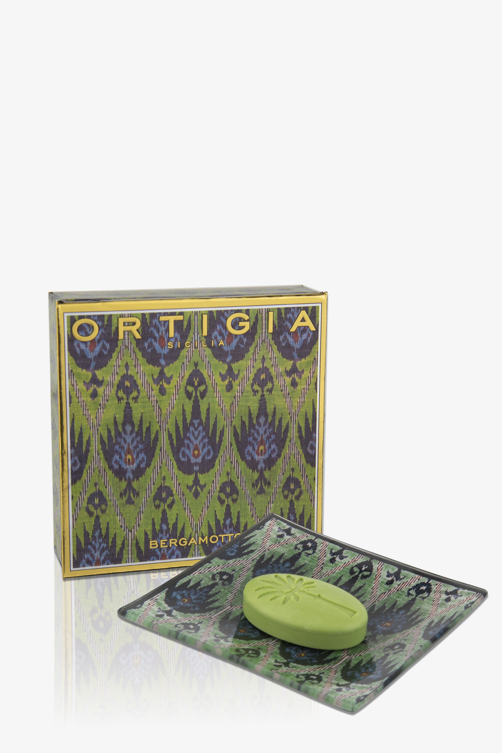 A green Ortigia Bergamot Soap Plate rests on a decorative glass dish in front of a patterned box with blue and gold accents.