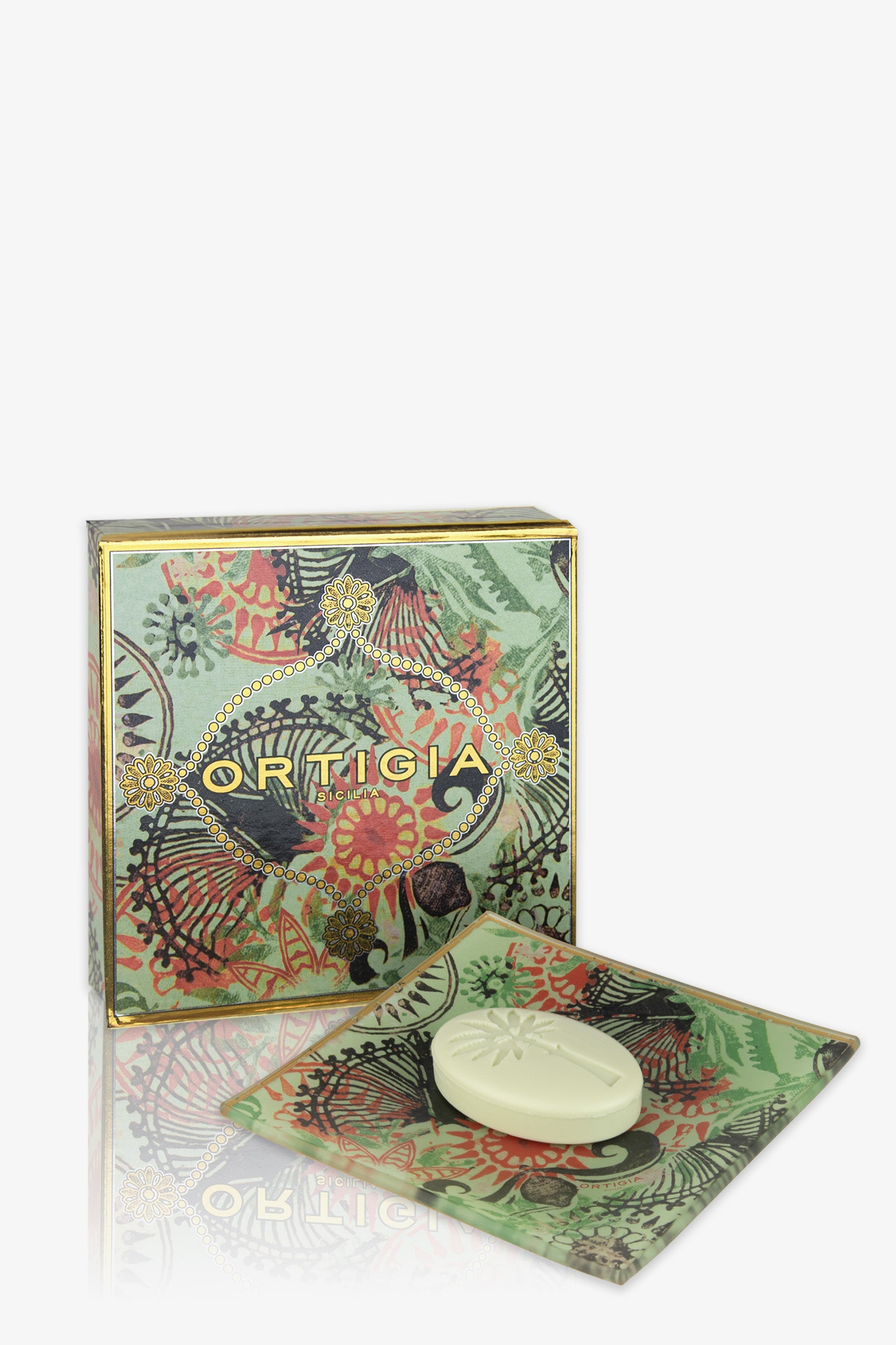 The Ortigia Glass Plate & Soap set features a decorative box with green, red, and gold botanical patterns, displayed behind a matching glass plate holding an oval embossed white soap.