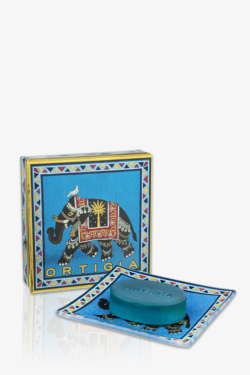 The Ortigia Sandalo Soap Plate Set in blue features a soap bar on a matching decorative tray, with a blue box adorned by a colorful, ornate elephant illustration in the background.