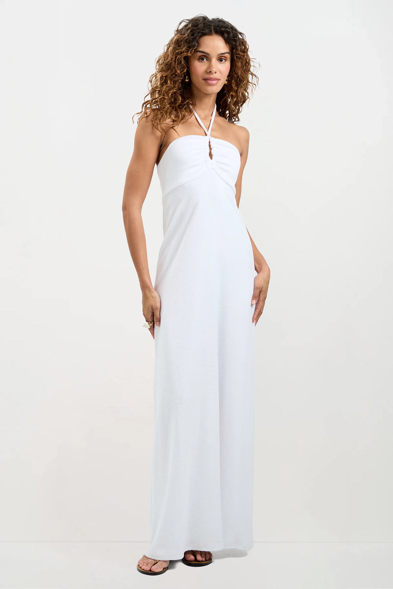 A woman with curly hair wears the Palm Sleeveless Maxi Dress - Poolside Terry, a strapless, floor-length white dress with halter tie detail and sandals. She stands against a plain white background, looking at the camera with a relaxed expression.