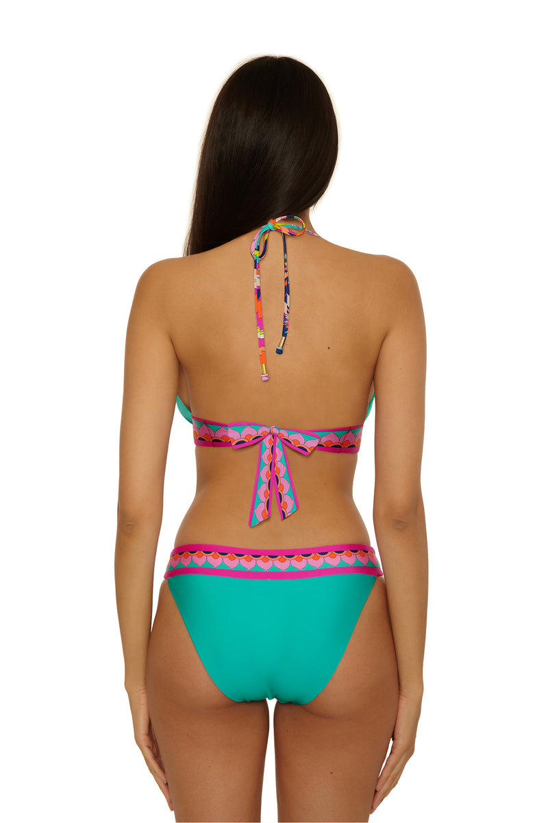 A woman with long brown hair stands against a white background, wearing the Paralia Reversible Hipster bikini featuring turquoise fabric and colorful geometric-patterned straps and waist.