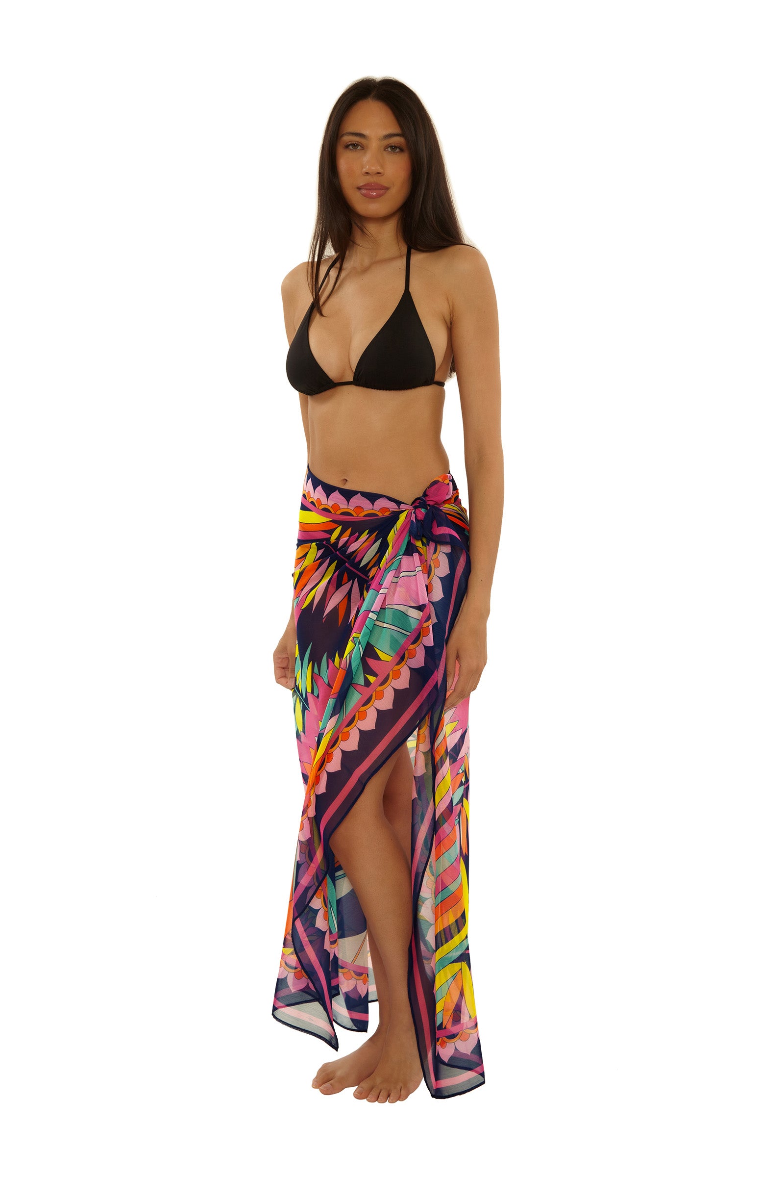 A woman wearing a black bikini top and the Paralia Pareo, a colorful patterned sarong skirt, stands barefoot against a white background.