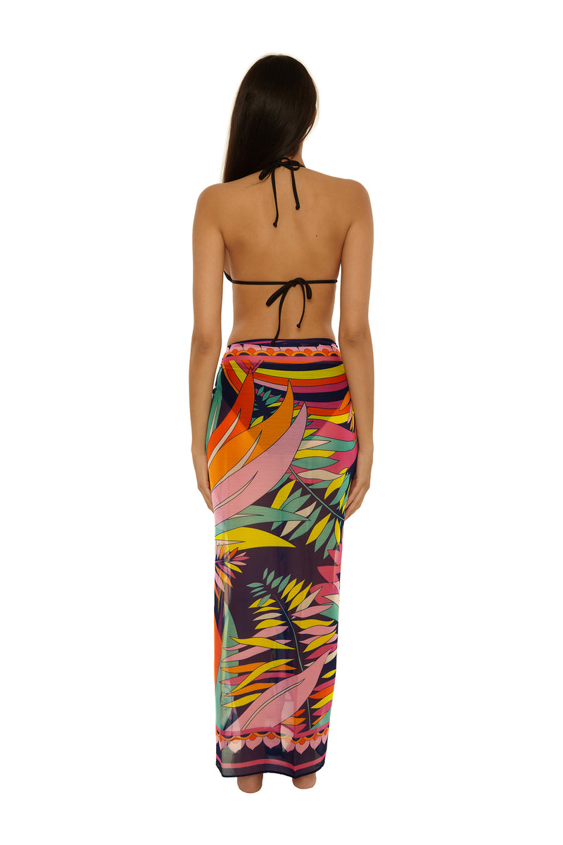 A woman with long brown hair models the Paralia Pareo, a colorful tropical-print sarong skirt, and a black halter bikini top, shown from the back against a white background.