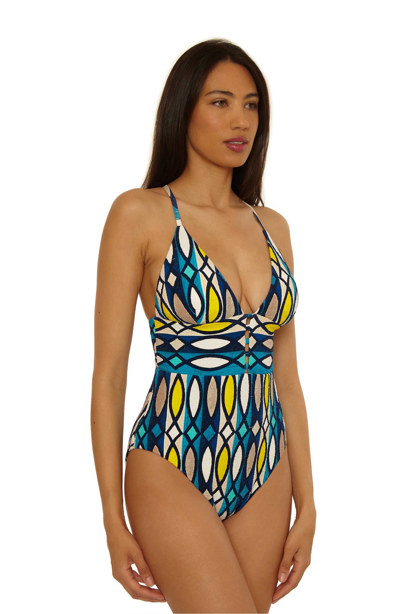 A woman with long dark hair wears the Paros Empire Waist Maillot, a colorful one-piece swimsuit with blue, yellow, white, and black geometric patterns. She stands slightly turned left against a white background.