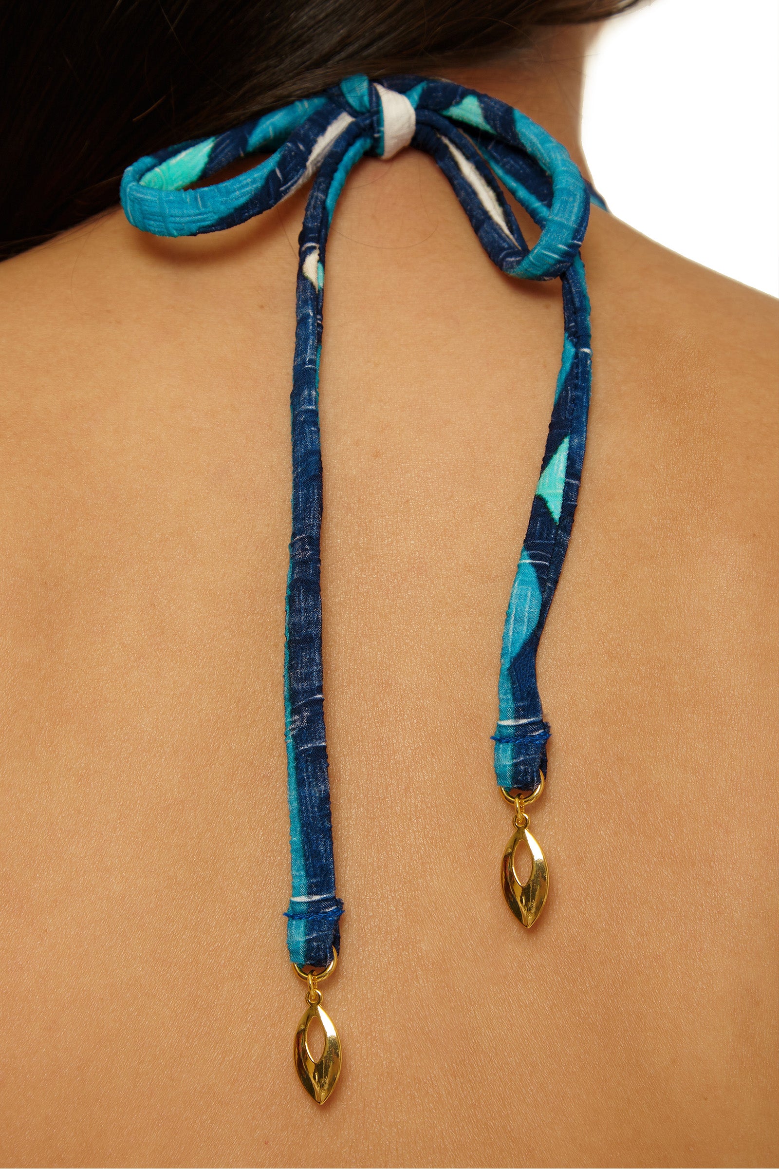 Close-up of a person’s upper back wearing the Paros Banded Bandeau Top, featuring blue patterned bikini strings tied in a bow with gold decorative charms; strands of the person's hair are partially visible at the top.