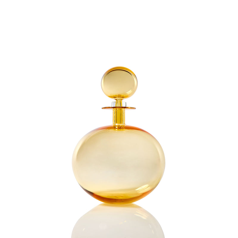 The Petite Low Round Decanter in whiskey, featuring an elegant silhouette and matching spherical cap, is showcased against a white background with a subtle reflection below.