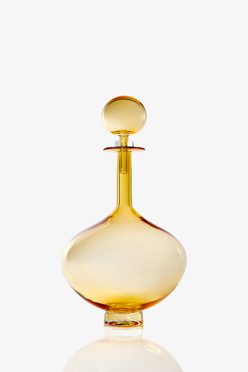 The Petite Genie Bottle Decanter features translucent amber glass with a round stopper, wide body, and short neck. It stands on a clear flat base and is set against a plain white background.