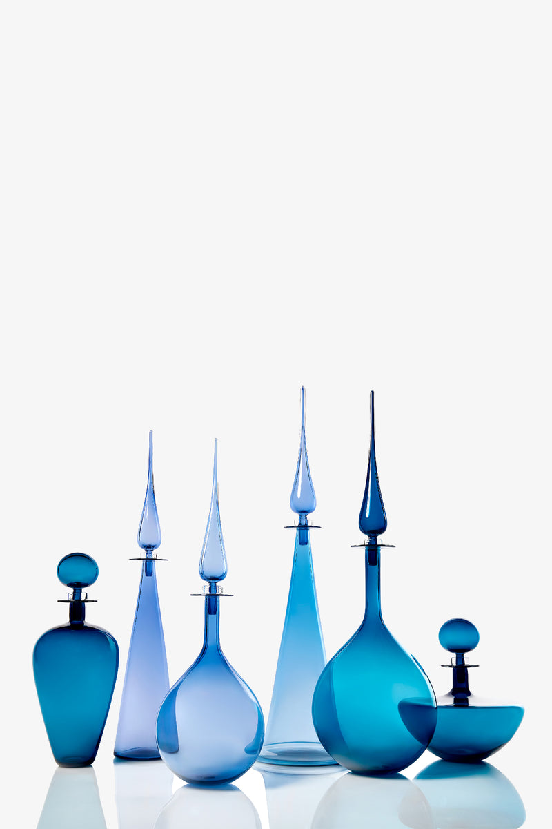 The Petite Straight Cone Decanter is showcased among six elegant blue glass bottles and decanters of different shapes and heights, each with a unique stopper, arranged on a reflective white surface against a plain white background.