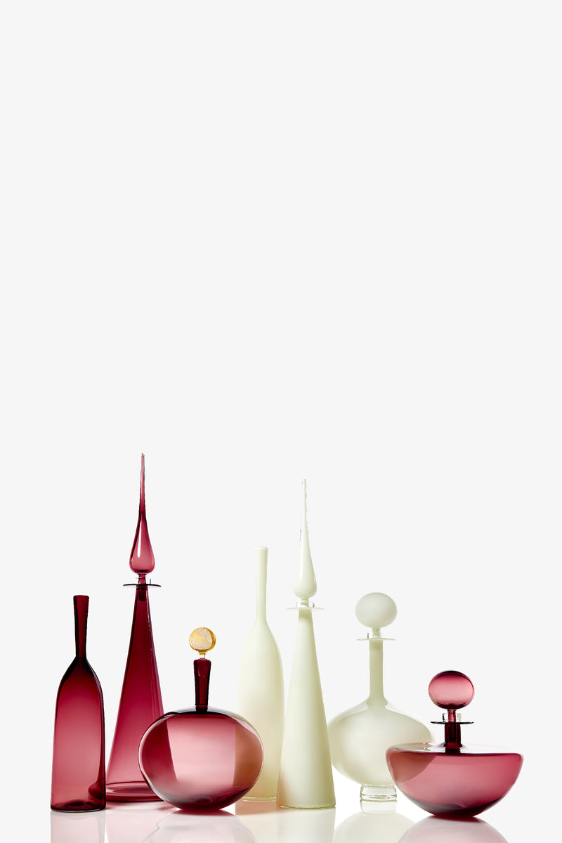 The Petite Straight Cone Decanter, an elegant glass bottle with a tall, slender shape and a decorative stopper, is displayed on a reflective surface against a white background.