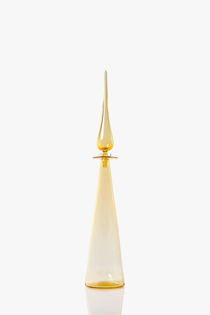 The Petite Straight Cone Decanter is a tall, elegant glass bottle with a pointed stopper in translucent amber-yellow, set against a plain white background.