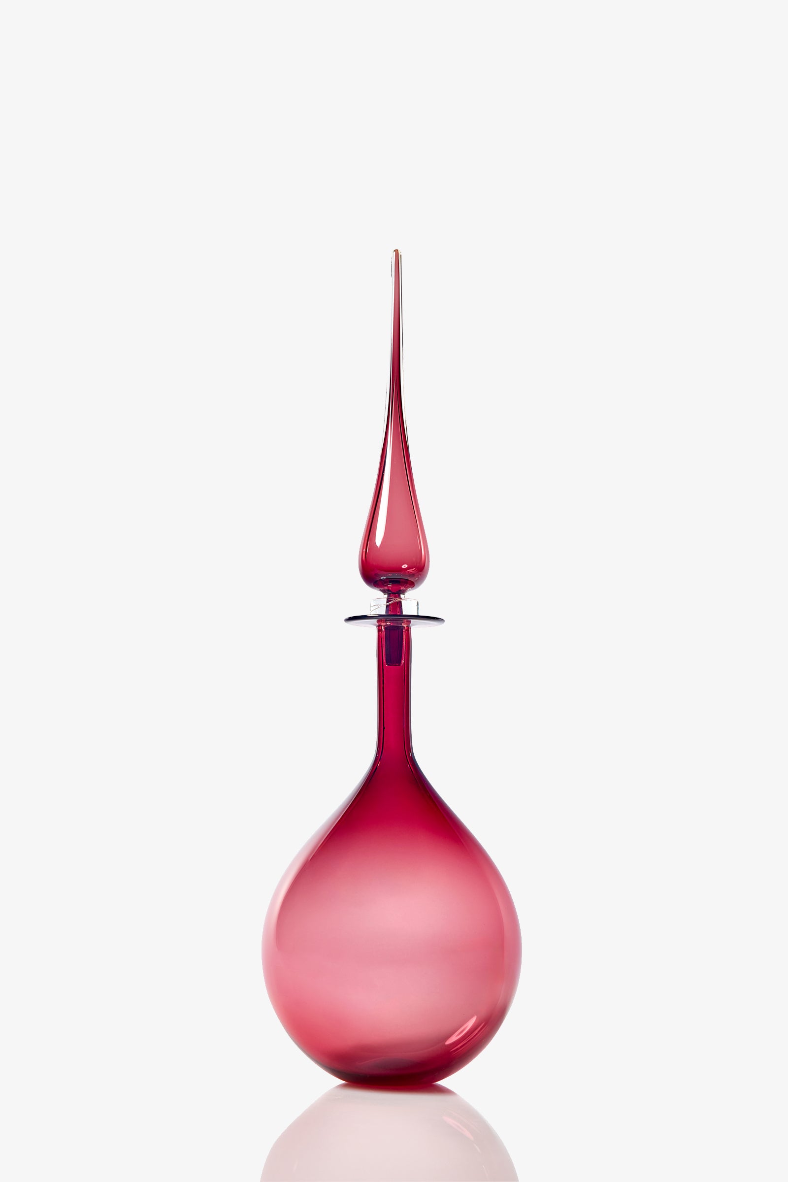 The Petite Tear Drop Decanter is a tall, elegant vessel with a round base and a long, pointed stopper, crafted from translucent pink glass and shown against a plain white background.