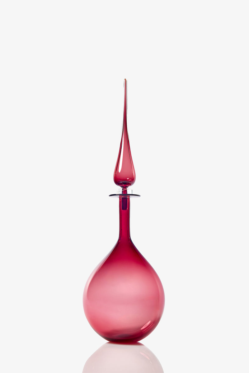 The Petite Tear Drop Decanter is a tall, elegant vessel with a round base and a long, pointed stopper, crafted from translucent pink glass and shown against a plain white background.