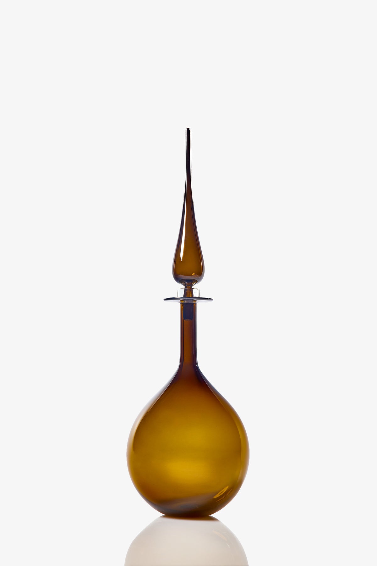 The Petite Tear Drop Decanter, a brown glass bottle with a round base and tall, slender pointed stopper, is photographed against a white background with a subtle reflection below.