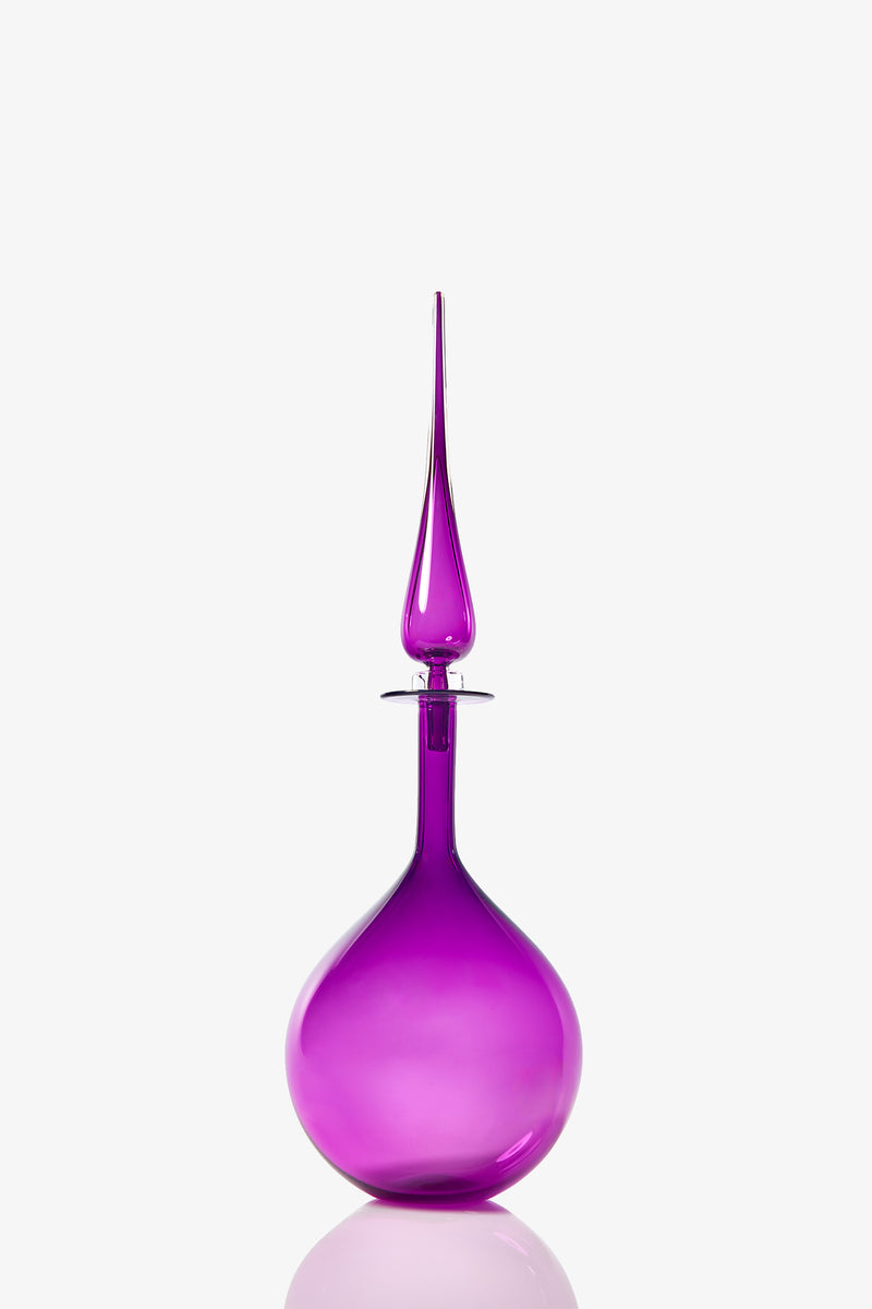 The Petite Tear Drop Decanter is a tall, elegant glass bottle with a bright purple hue, a round base, and a long pointed stopper, shown against a plain white background.