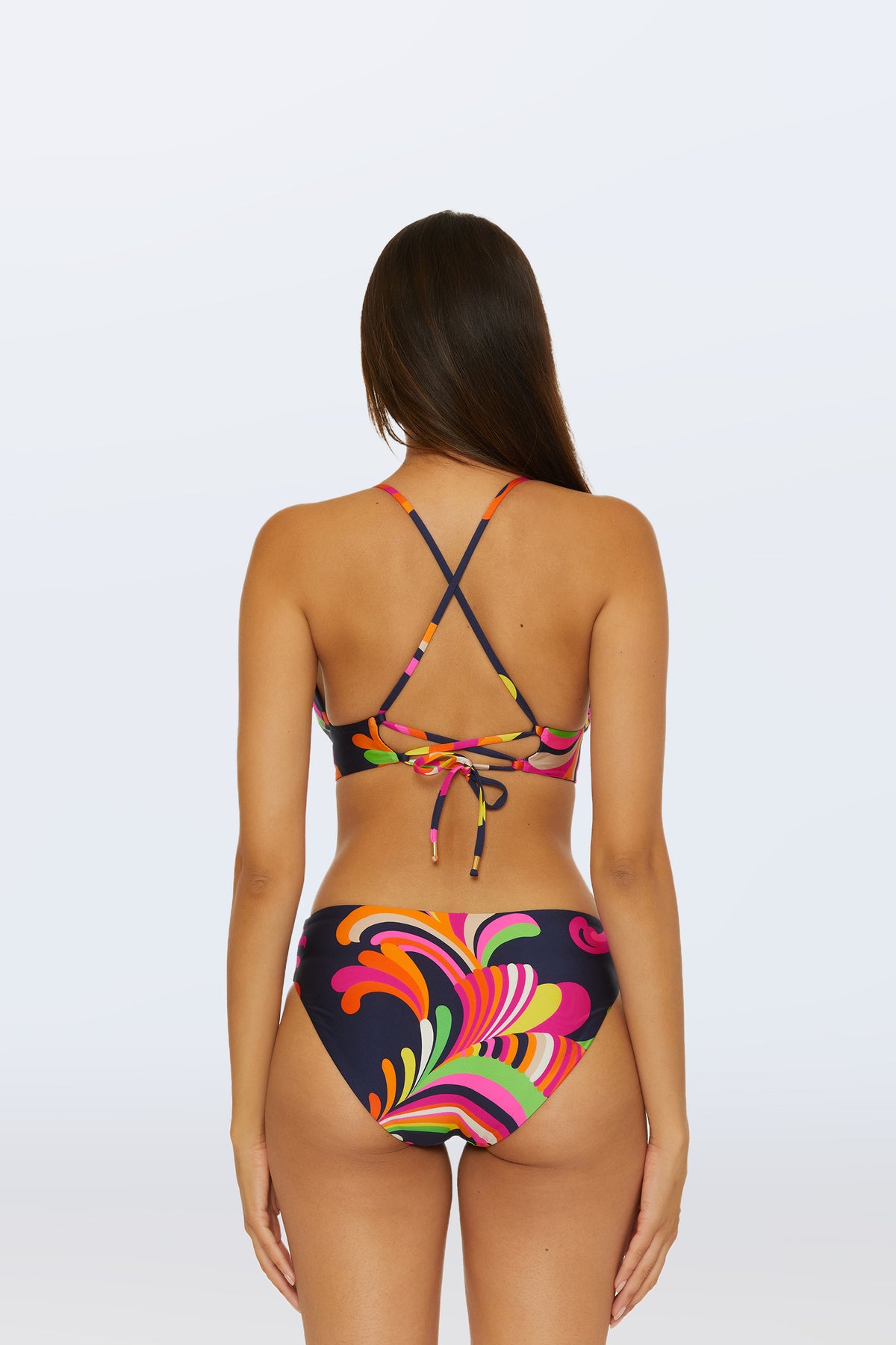 A woman with long brown hair is shown from the back wearing the Reversible Halter Bra Swim Top - Palms Paradise, featuring crisscross straps and a tie back with vibrant orange, pink, yellow, and green swirls on a dark background.