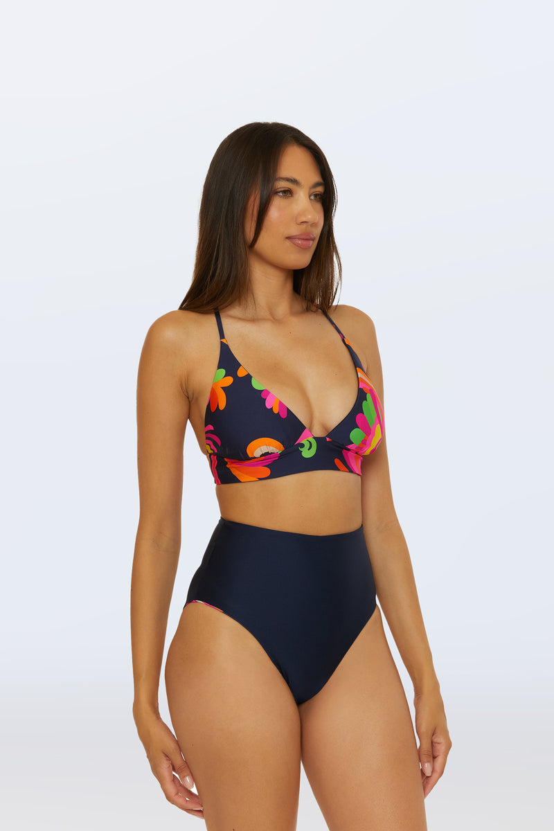 A woman with long brown hair wears a dark floral bikini top and the Reversible High Waist Bottom - Palms Paradise, standing against a plain light background.