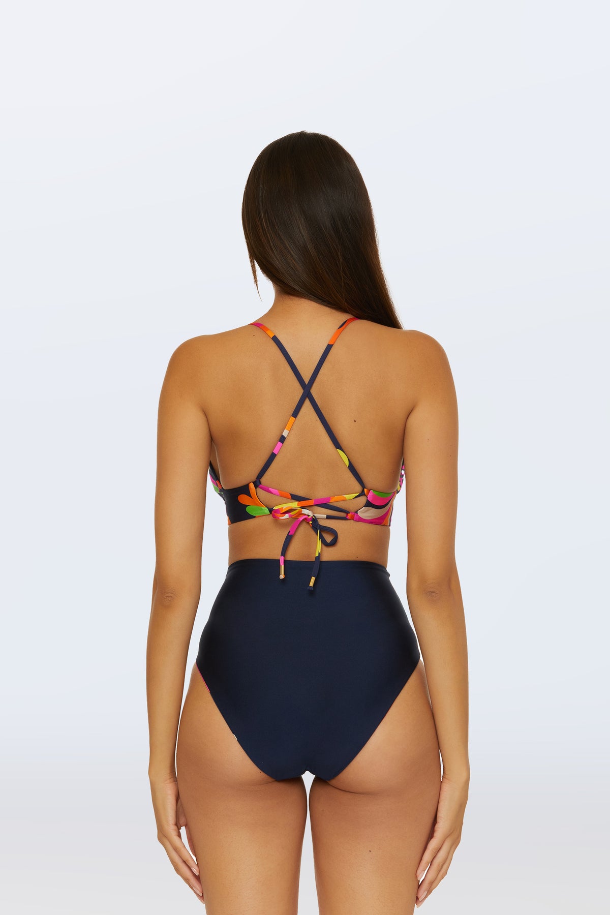 A woman with long brown hair stands against a light background, wearing the Reversible High Waist Bottom - Palms Paradise in navy and a colorful bikini top with crisscross straps and a tie at the back.