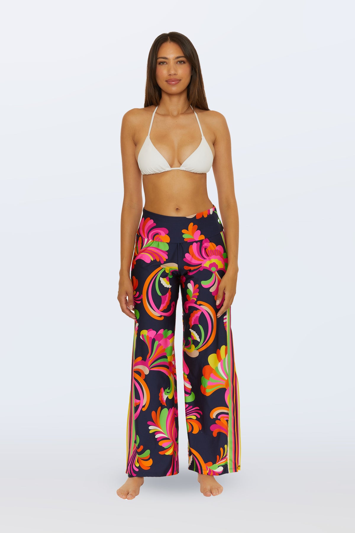 A woman stands barefoot facing forward against a plain light backdrop, wearing a white bikini top and the Swim Cover-Up Pant - Palms Paradise in Multi, featuring high-waisted, wide-leg design with a bold floral pattern.