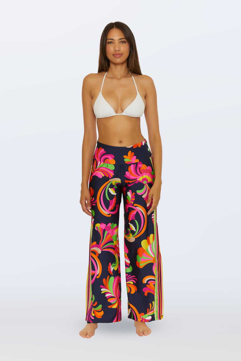 A woman stands barefoot facing forward against a plain light backdrop, wearing a white bikini top and the Swim Cover-Up Pant - Palms Paradise in Multi, featuring high-waisted, wide-leg design with a bold floral pattern.