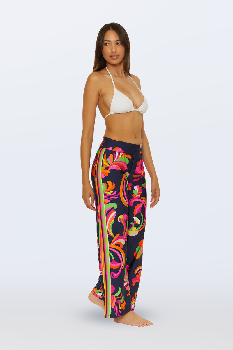 A woman stands barefoot in a white bikini top and the Swim Cover-Up Pant - Palms Paradise, featuring high-waisted, loose-fitting navy pants with a colorful floral and swirl pattern plus striped sides, against a plain light background.