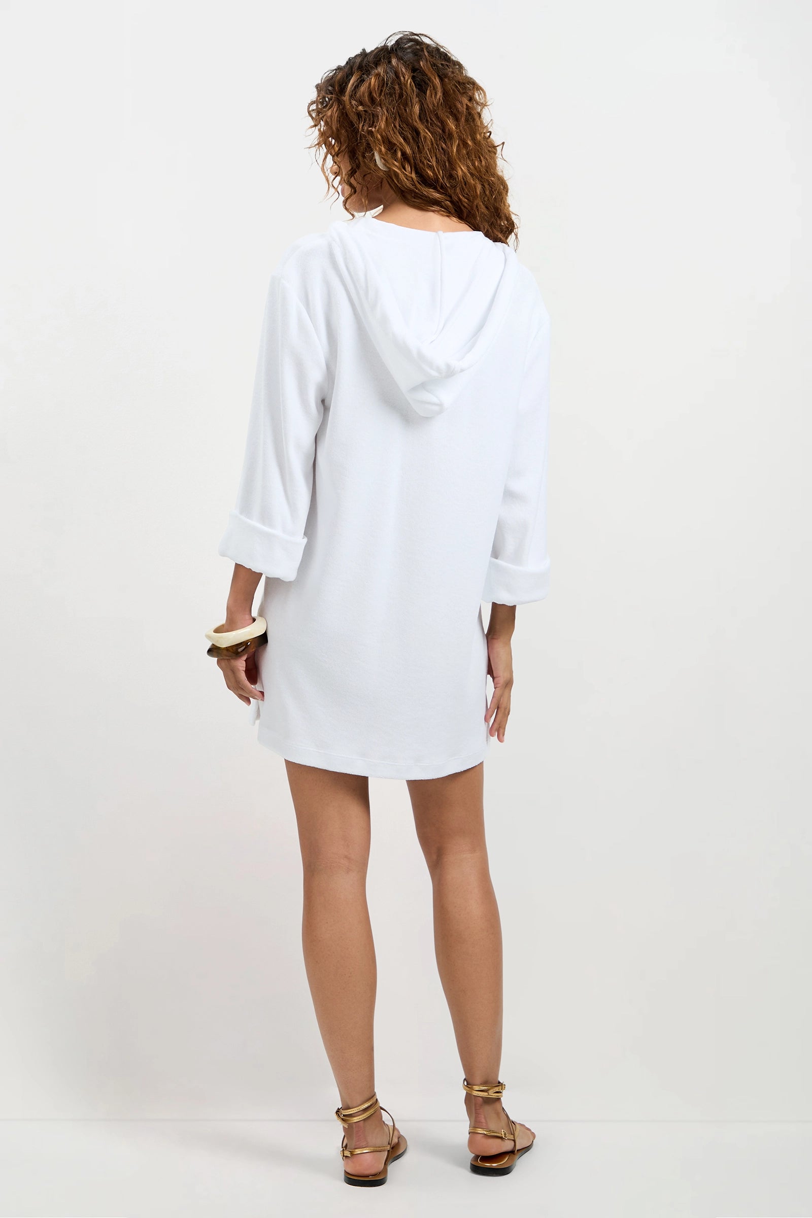 A woman with curly hair stands facing away, wearing the Poolside 3/4-Sleeve Hoodie in white terry, styled as a dress with rolled-up sleeves, gold bracelets, and sandals against a plain white background.