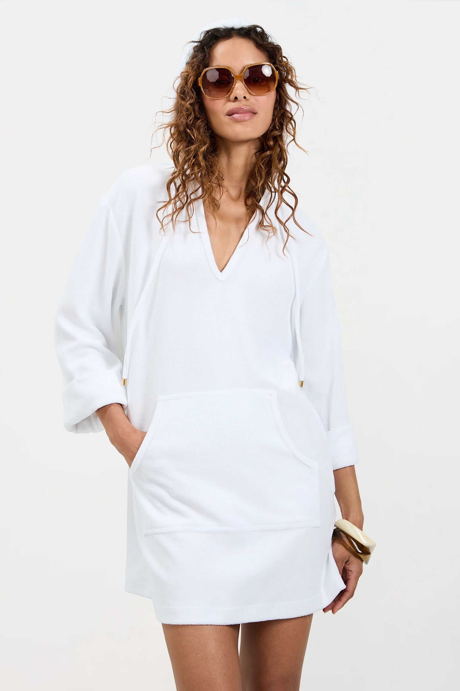 A woman with curly hair and sunglasses stands confidently against a plain background, wearing the Poolside 3/4-Sleeve Hoodie in Poolside Terry and holding a light-colored clutch.