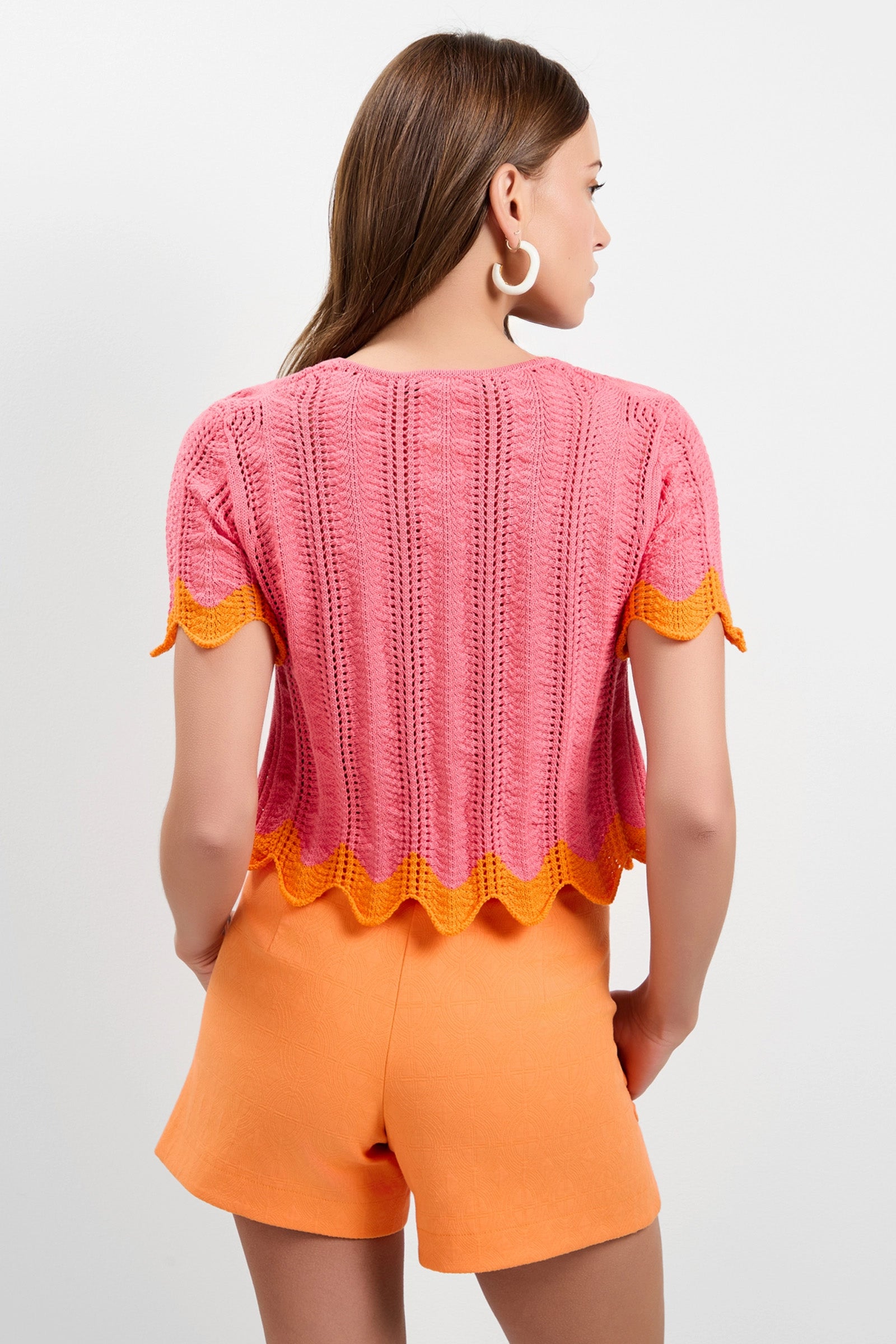 A woman with long brown hair and large white hoop earrings wears the Playa Largo Wavy Knit Top—pink, short-sleeve, with orange scalloped trim—paired with high-waisted orange shorts, standing with her back to the camera against a white background.