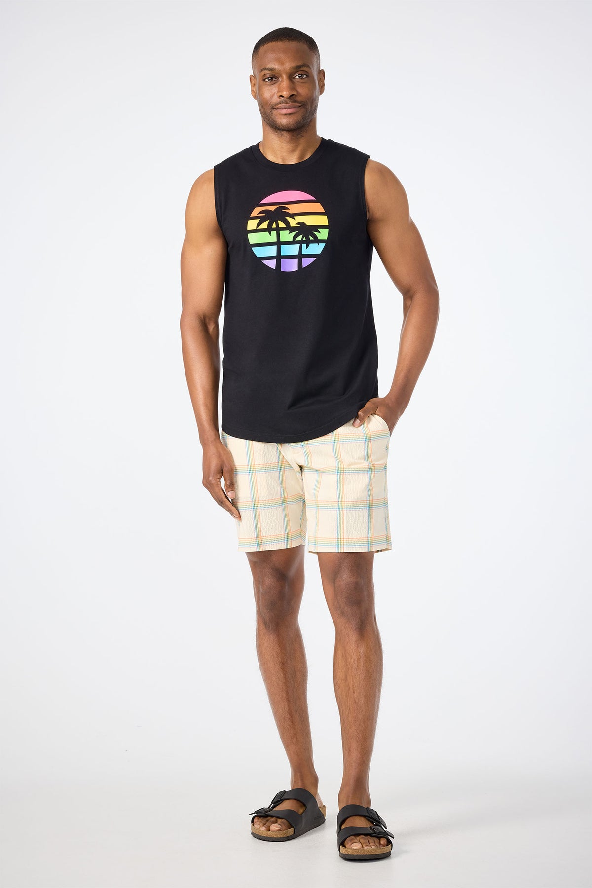 A man stands against a plain background wearing the Sleeveless Muscle Tee - Pride, featuring a rainbow sunset and palm trees graphic. He pairs it with plaid shorts and black sandals, with one hand in his pocket and a slight smile.