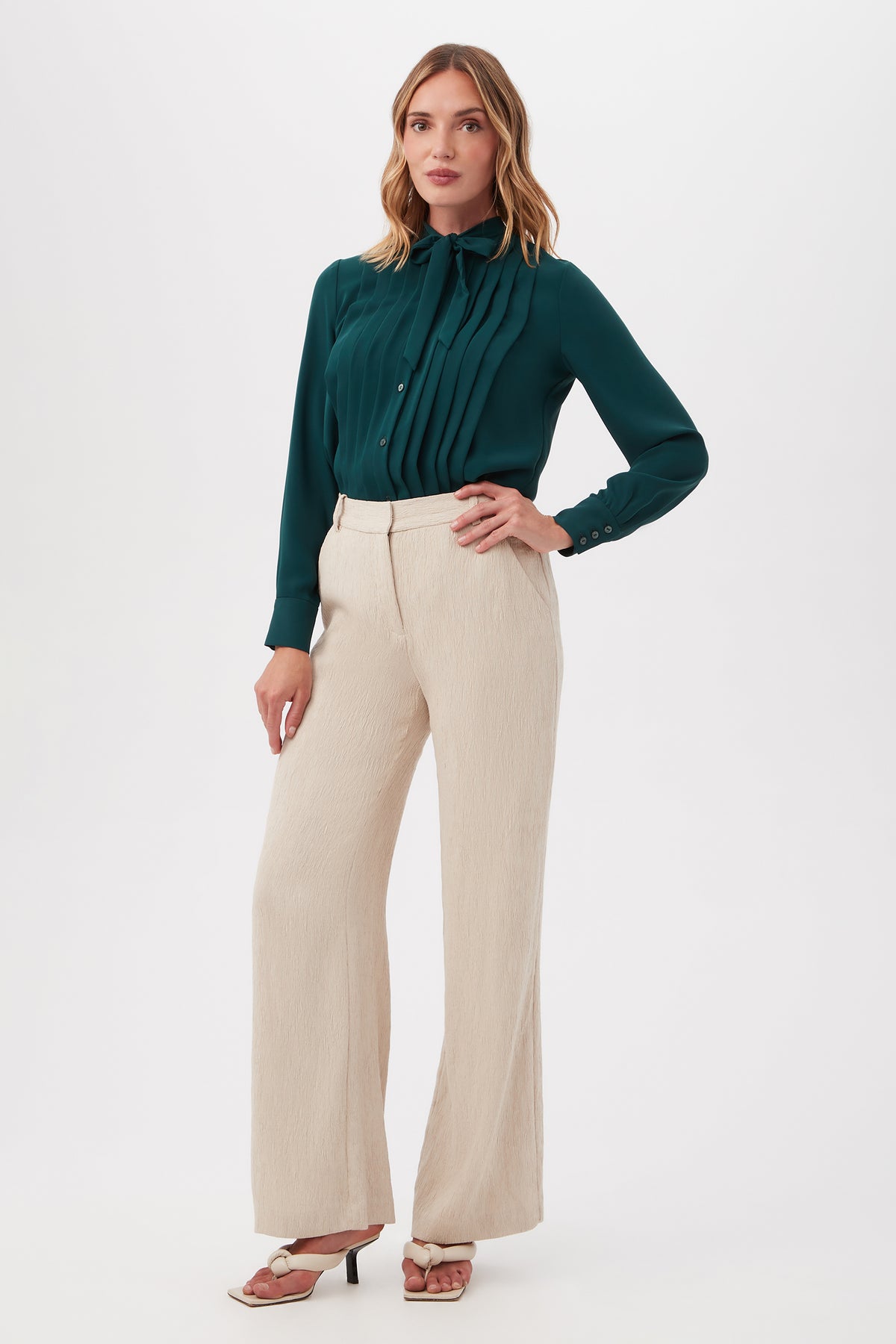 A woman stands against a white background wearing the Kahina Pant, cream-colored and wide-leg, paired with a dark green pleated blouse. She has loose, wavy hair and completes her look with heeled sandals.