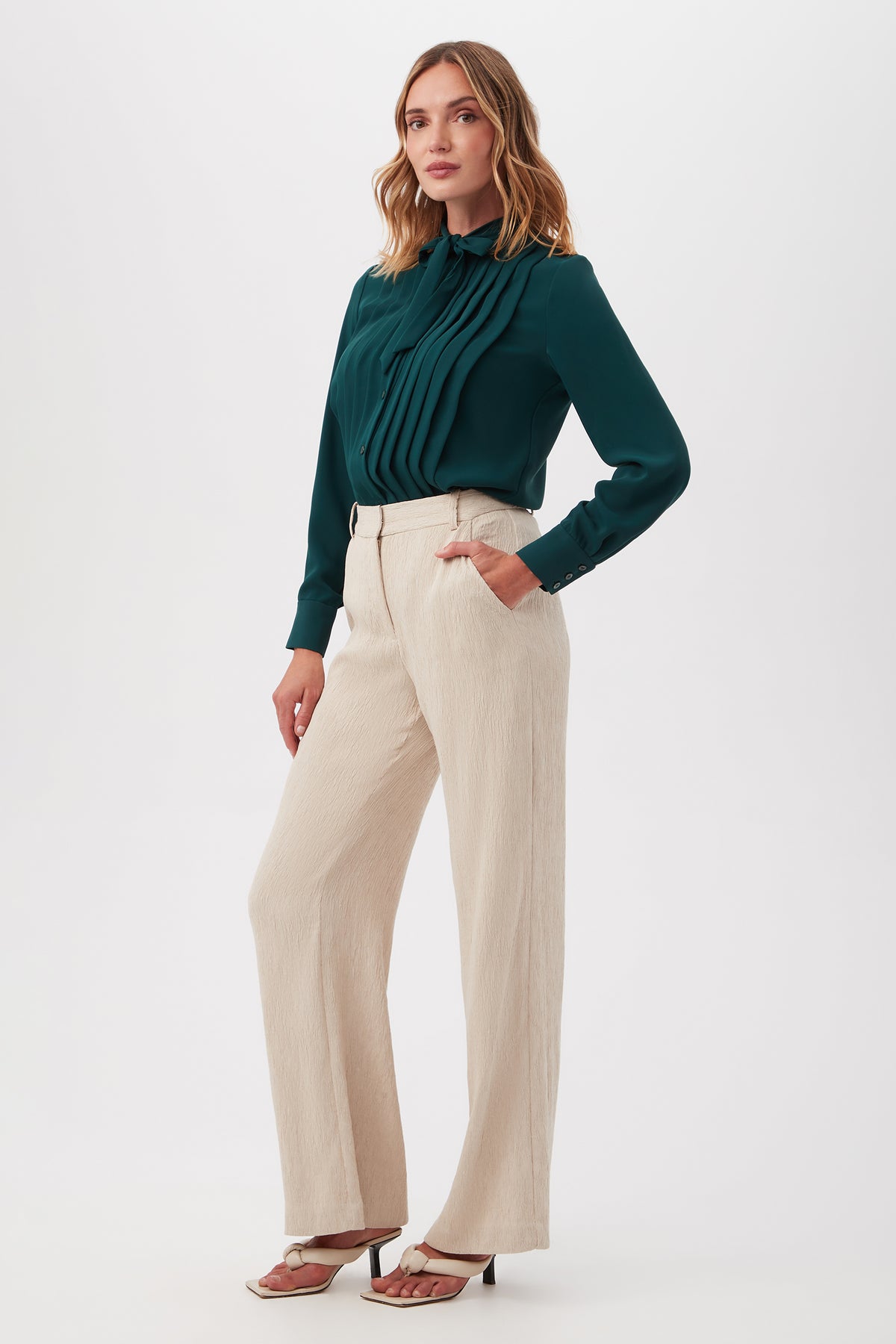 Woman stands against a plain background in beige high-waisted Kahina Pant, a dark green pleated long-sleeve blouse, and white heeled sandals. She has wavy shoulder-length hair and poses casually.