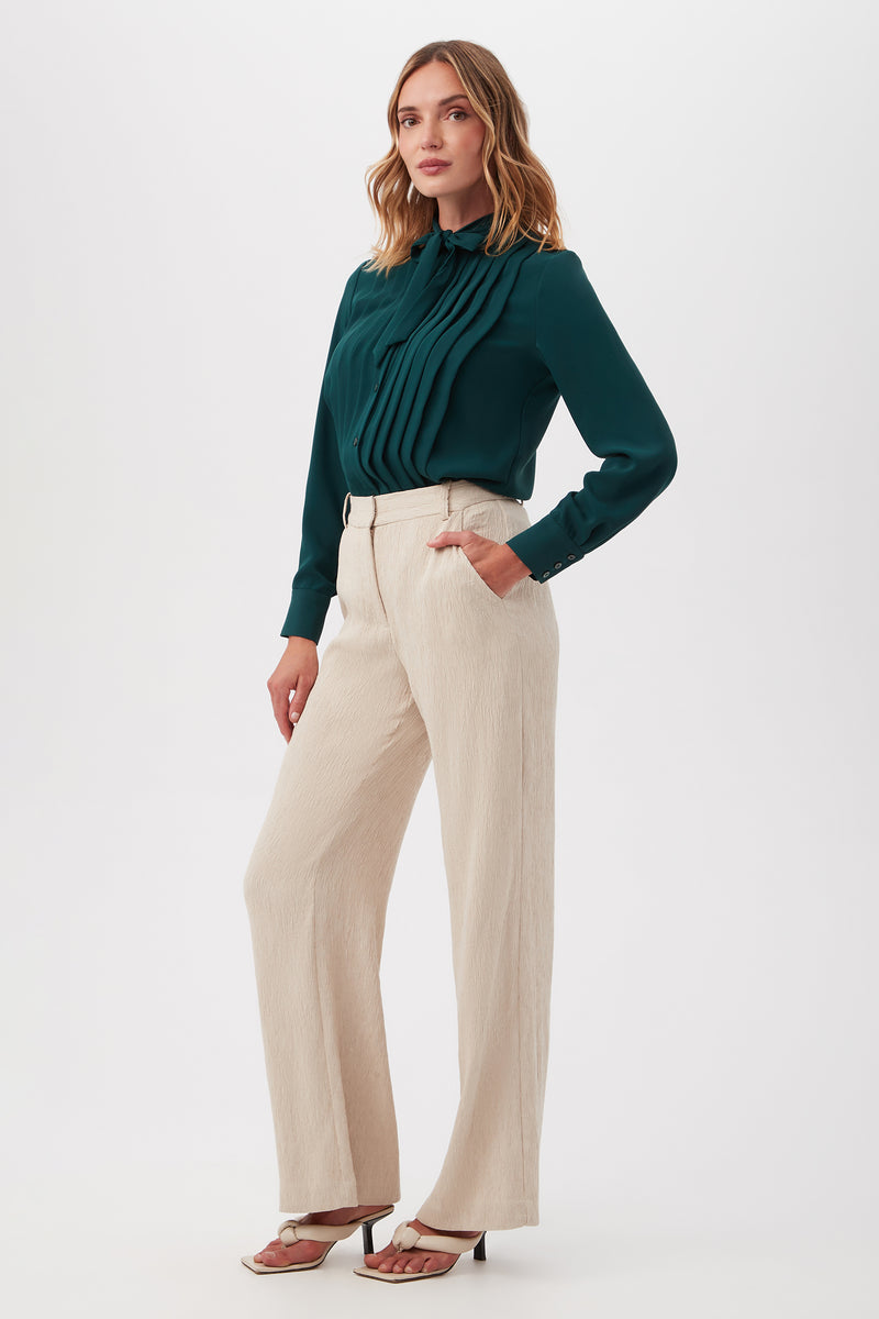 Woman stands against a plain background in beige high-waisted Kahina Pant, a dark green pleated long-sleeve blouse, and white heeled sandals. She has wavy shoulder-length hair and poses casually.