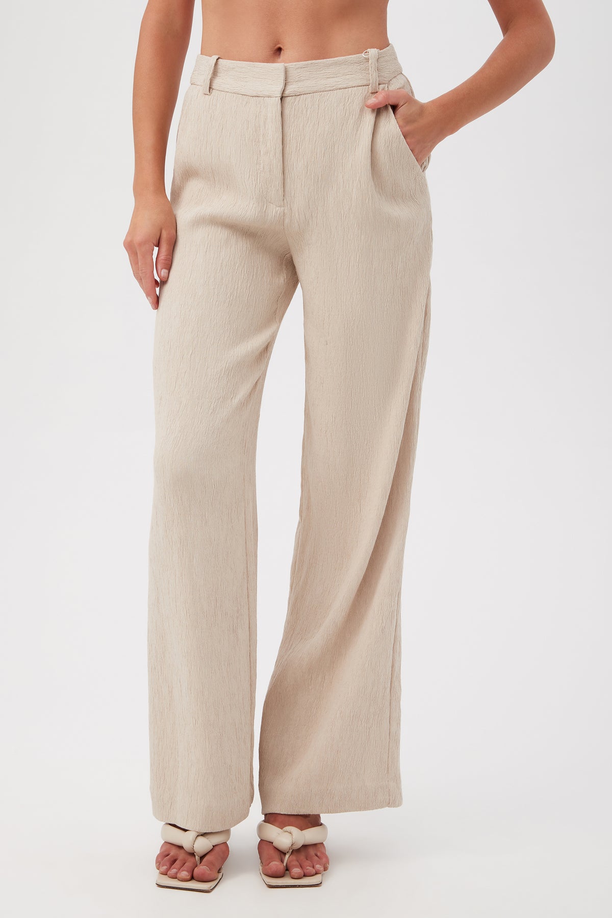 A person wearing the Kahina Pant in Sahara Stone, styled with cream braided sandals and one hand in a pocket, stands against a plain white background.