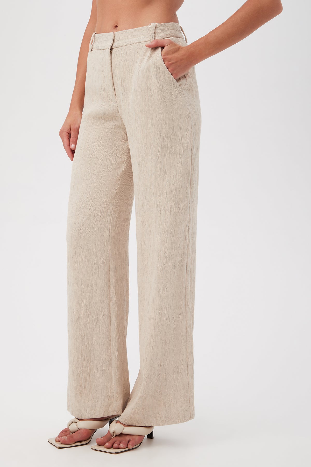 A model showcases the Kahina Pantâ€”high-waisted, wide-leg, beige textured pants with front pocketsâ€”paired with heeled sandals against a plain white background. Only the lower torso and legs are visible.