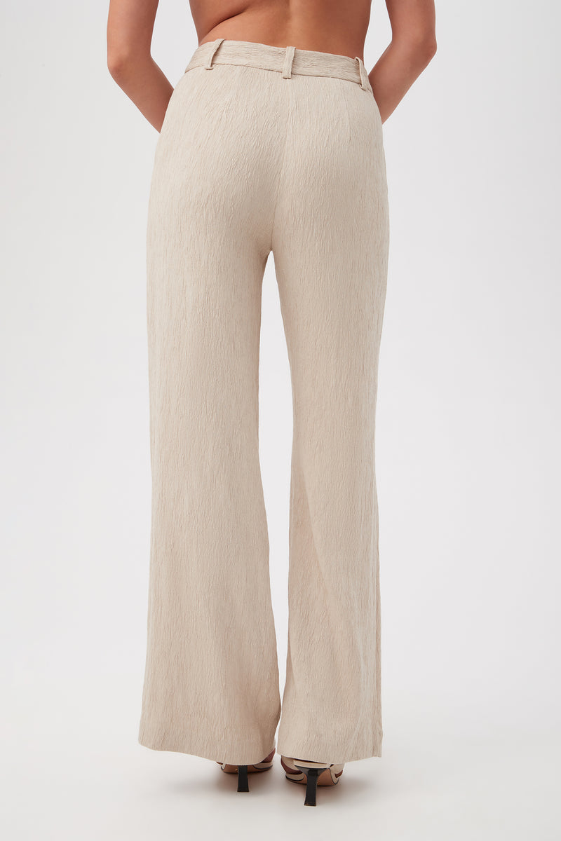 A person is shown from the waist down wearing the Kahina Pantâ€”high-waisted, wide-leg, textured beige trousersâ€”and heeled sandals, standing against a plain light background.