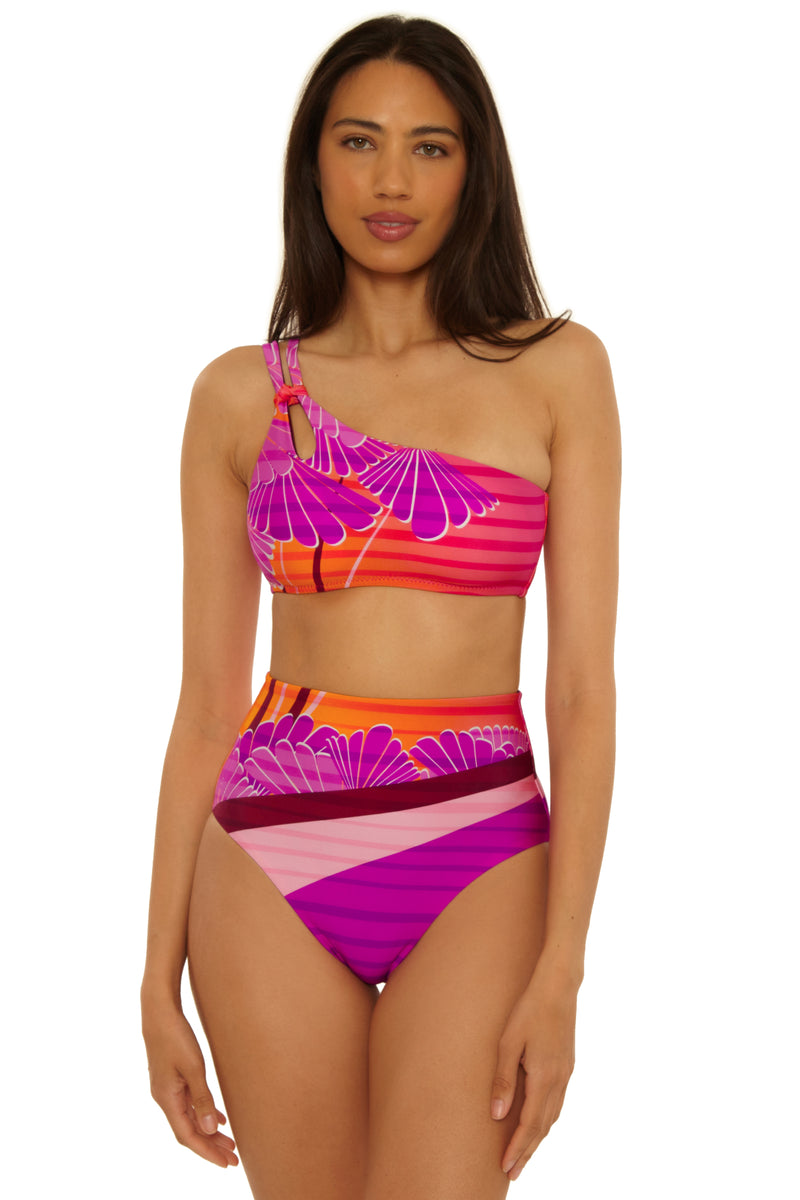 A woman models the Rhodes One Shoulder Top bikini, featuring a retro pattern in pink, purple, orange, and red hues with a ring detail and high-waisted bottoms. She faces forward with a neutral expression.