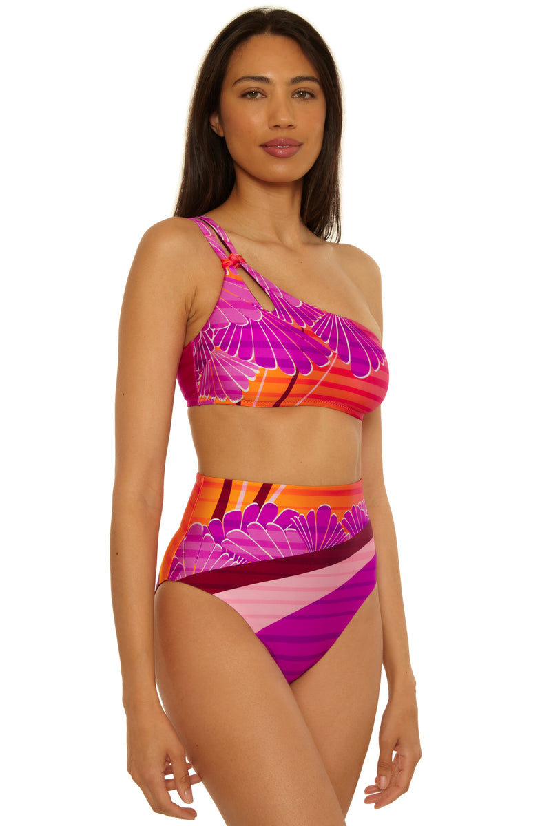 A woman wears the Rhodes One Shoulder Top bikini featuring a bold pink, orange, and purple geometric pattern, standing facing forward and slightly to the side against a white background.