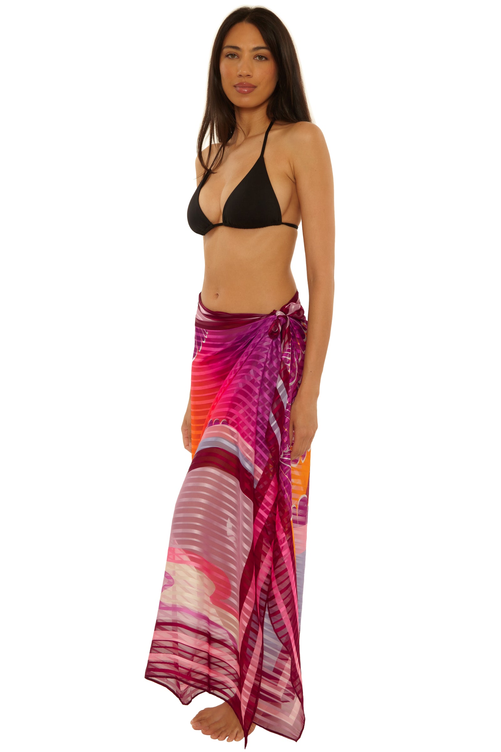 A woman wears the Rhodes Pareo, a colorful long sheer sarong in pink, purple, and orange shades, over a black bikini top while standing barefoot against a plain white background.