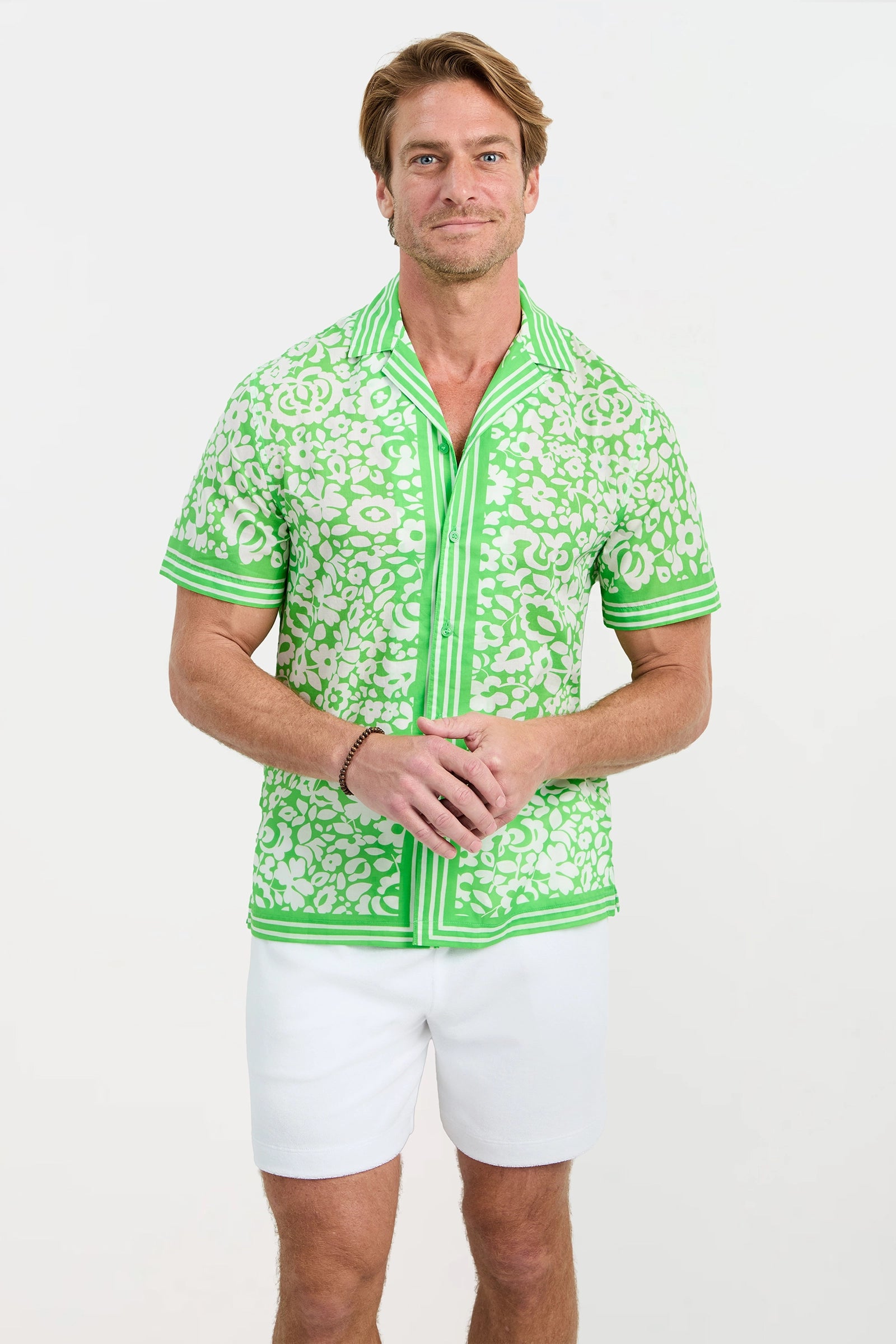 A man with light brown hair wears the Josue Short-Sleeve Shirt - Poolside Gossip and white shorts, standing against a plain white background and smiling with his hands clasped.