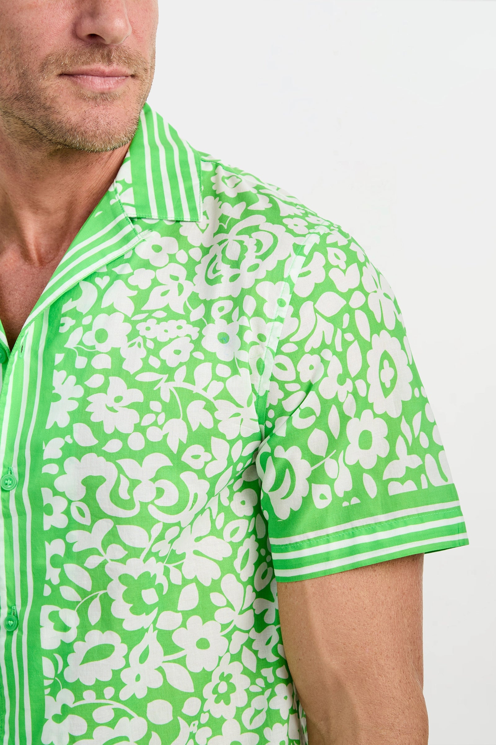A man wears the Josue Short-Sleeve Shirt - Poolside Gossip, featuring a green color with a white floral pattern and white stripes on the collar, placket, and sleeve cuffs. Only his lower face and upper torso are visible.