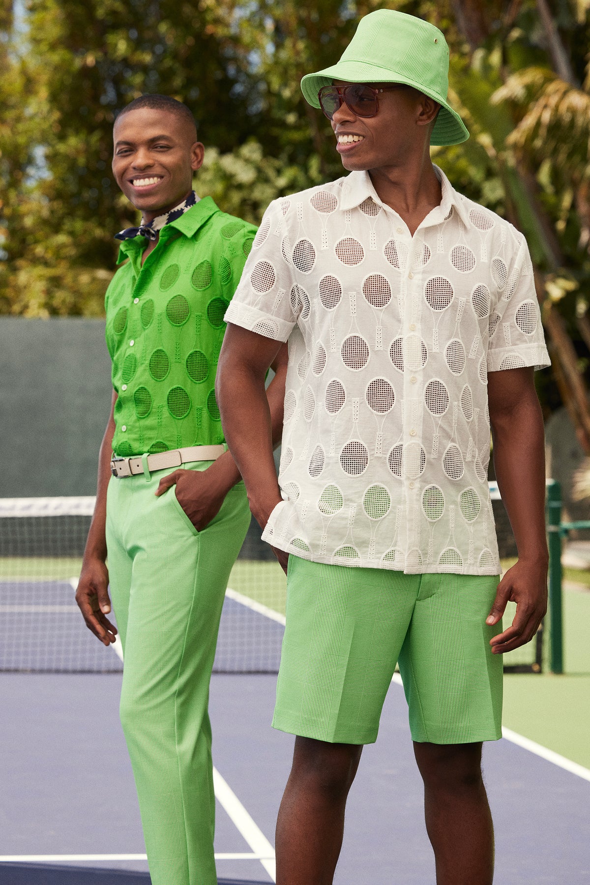Two men stand on a tennis court in green outfits with bold polka dots. One wears trousers, a belt, and the Branson Shirt - Tennis Eyelet; the other sports shorts, a sheer shirt, sunglasses, and a green bucket hat.