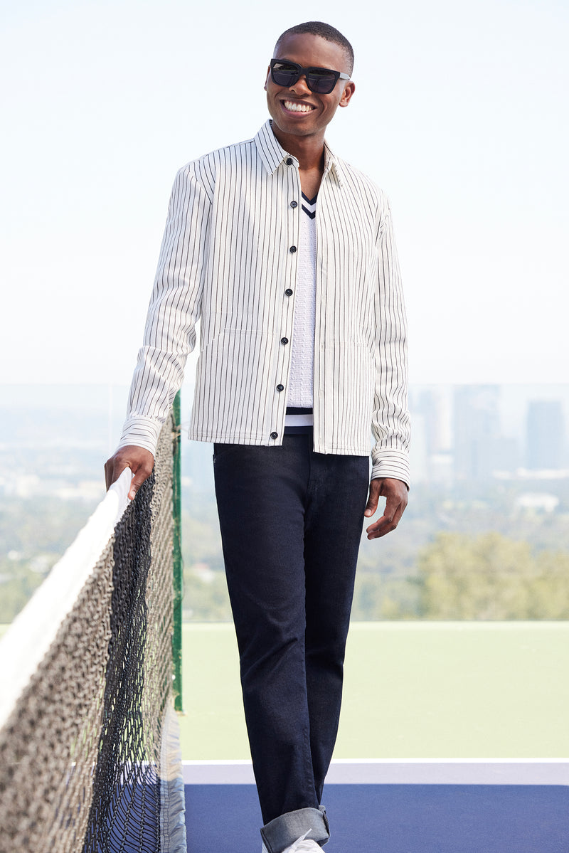 A smiling man in sunglasses and the Mulholland Shirt Jacket - Seles Stripe stands by a tennis net on an outdoor court, cityscape in the background.