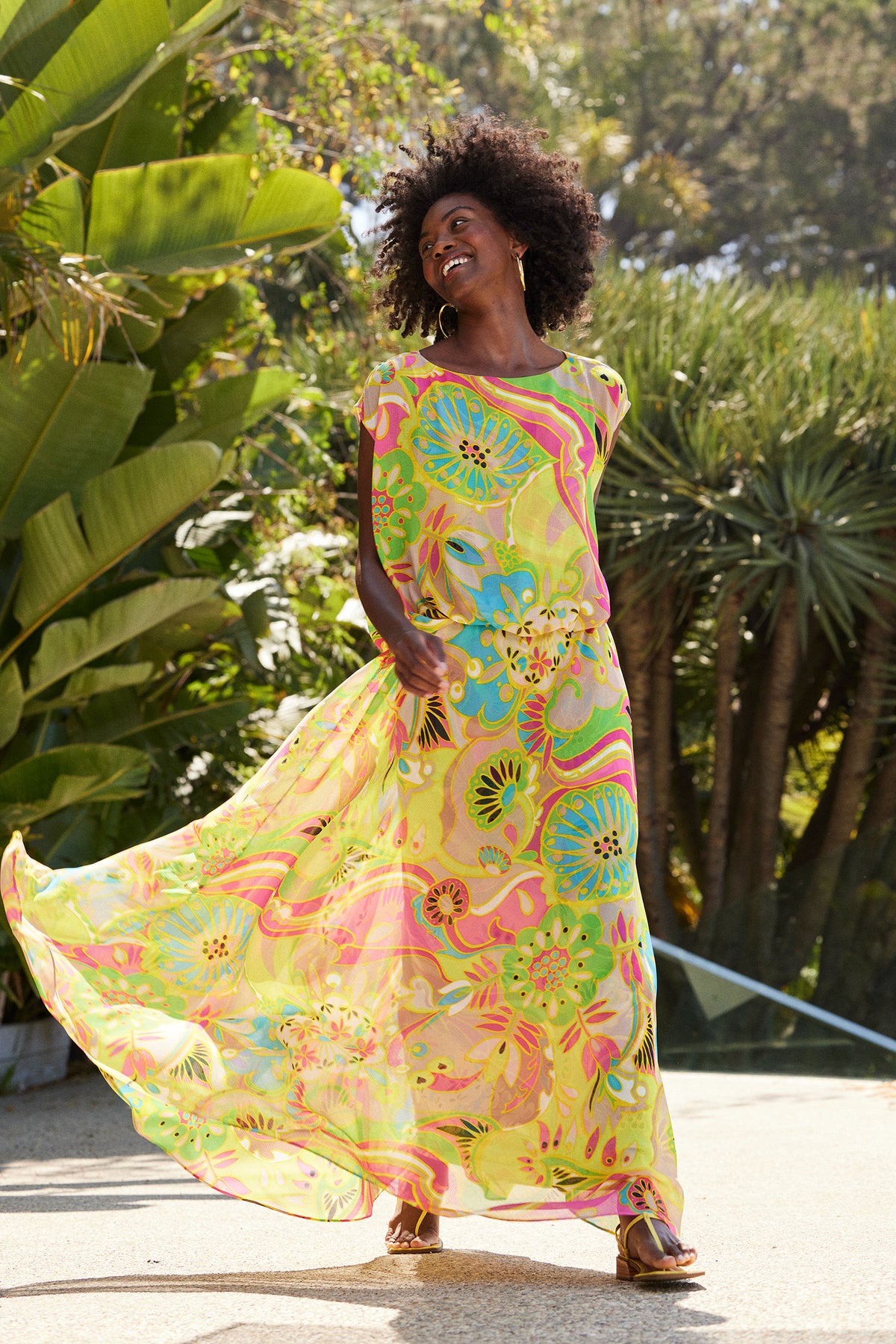 A woman smiles outdoors in bright sunlight, wearing the Vantage Short-Sleeve Maxi Dress - Venus with flowing, colorful floral and abstract patterns, surrounded by lush green plants.
