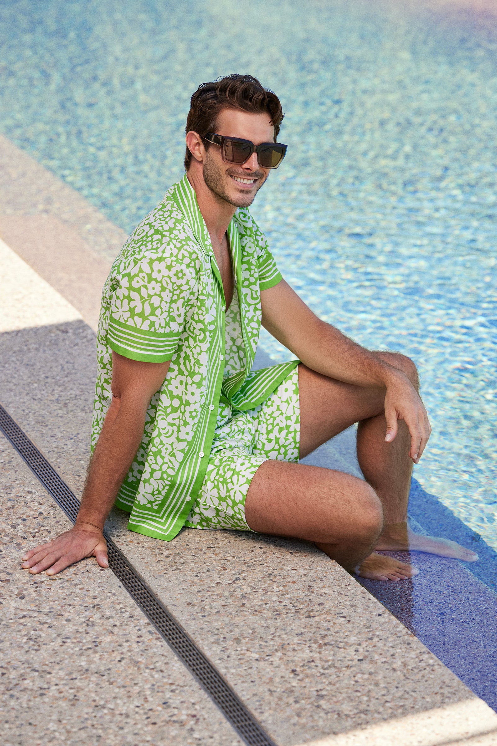 A man sits poolside with his feet in the water, smiling in the Josue Short-Sleeve Shirt - Poolside Gossip, featuring a light green and white floral pattern. He pairs it with matching shorts and large dark sunglasses on a bright sunny day.