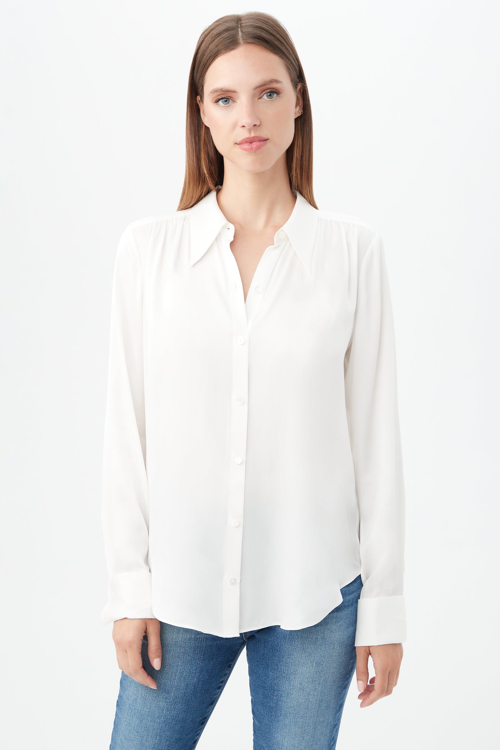 A woman with straight brown hair wears the Cassavetes Long-Sleeve Stretch Silk Button-Up Shirt in Whitewash and blue jeans, standing against a plain white background and looking at the camera with a neutral expression.