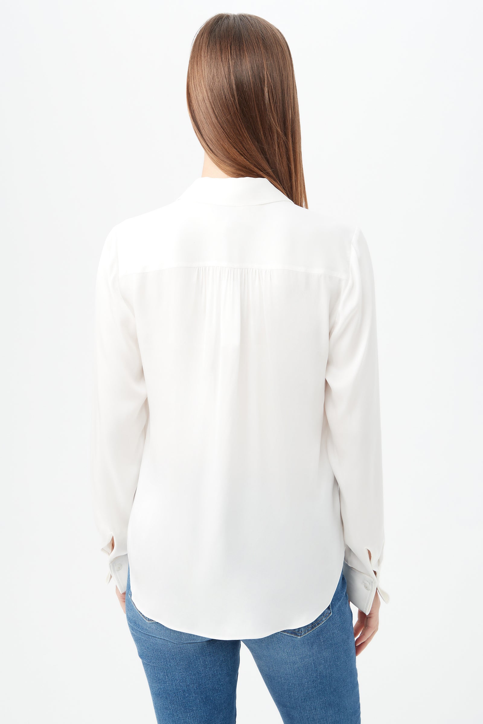 A woman with long brown hair wears the Cassavetes Long-Sleeve Stretch Silk Button-Up Shirt in white and blue jeans, standing with her back to the camera against a plain white background.