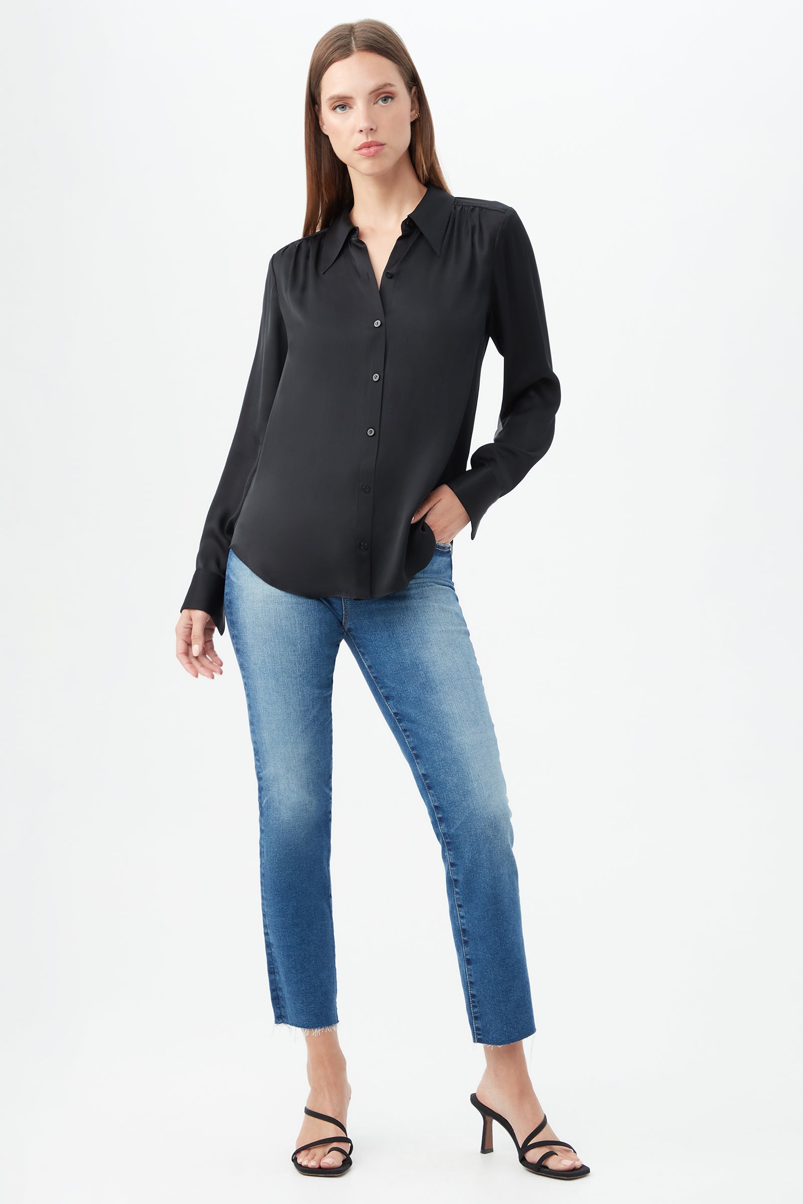 A woman stands against a plain white background wearing the Cassavetes Long-Sleeve Stretch Silk Button-Up Shirt in black, paired with light blue jeans and black heeled sandals. She poses with one hand in her pocket and a neutral expression.