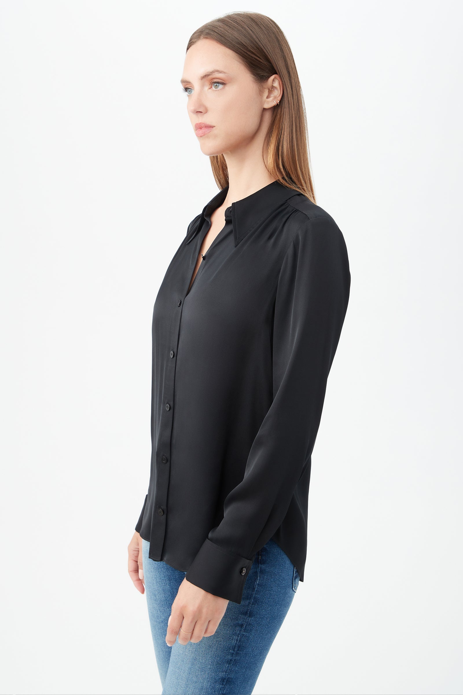A woman with straight, light brown hair wears the Cassavetes Long-Sleeve Stretch Silk Button-Up Shirt in black and blue jeans, standing sideways against a white background to showcase effortless women's fashion.
